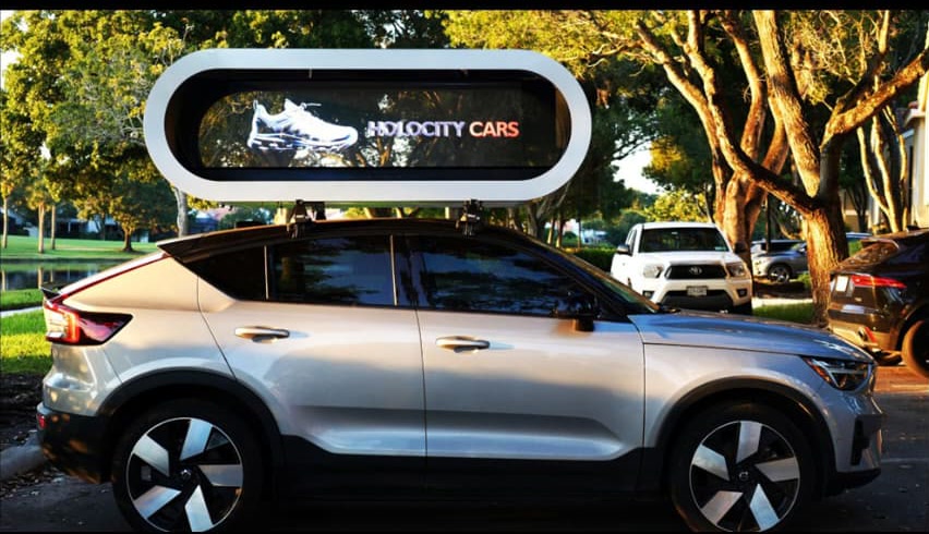 A silver Volvo crossover with a Holocity Cars hologram pod on its roof, showing a sneaker hologram in daylight
